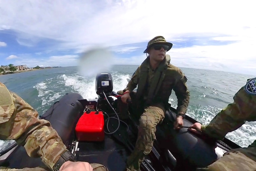 Ethan is standing on a black boat with a group of military soldiers sitting beside him