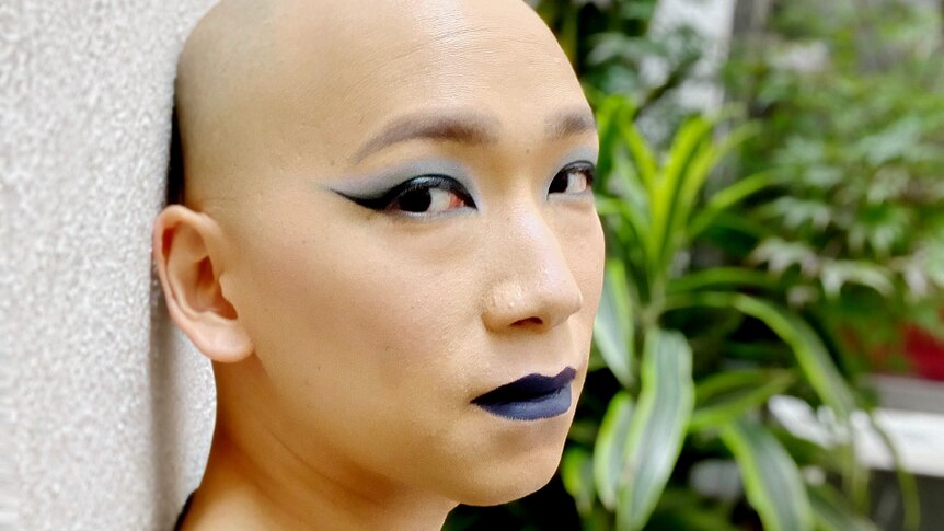 Person with bald head looking sideways at camera. Wearing blackish purple lipstick and blue eyeshadow
