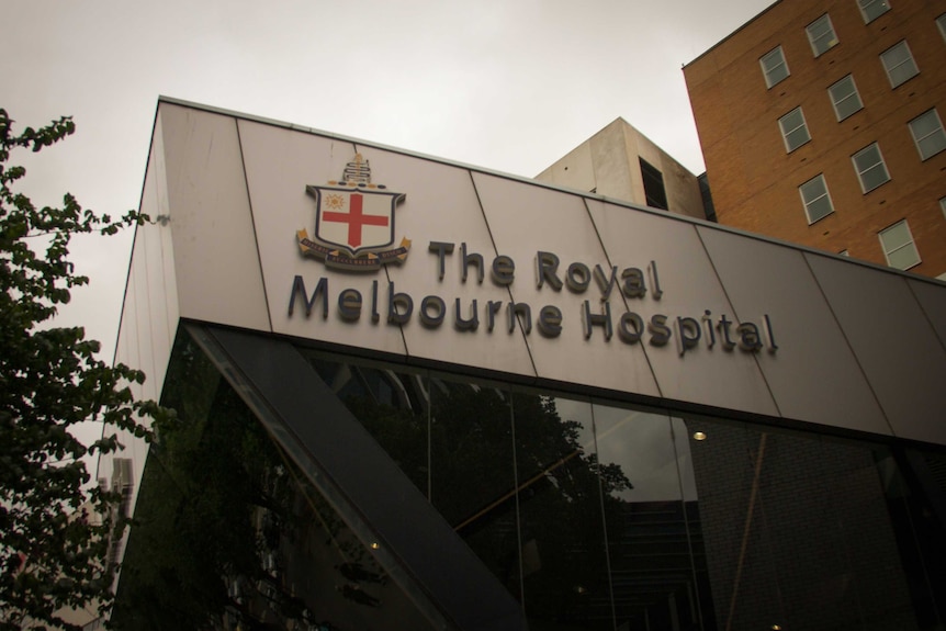A sign at the Royal Melbourne Hospital as a building with many windows towers in the background. 