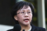 Madam Dong speaks at a media conference. She wears glasses, lipstick, a pearl necklace and black