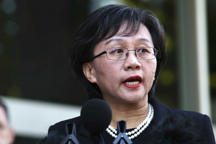 Madam Dong speaks at a media conference. She wears glasses, lipstick, a pearl necklace and black