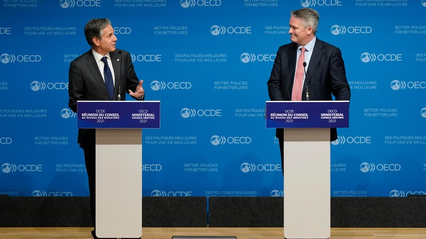 Antony Blinken and Mathias Corman stand on stage in front of OECD podiums.  