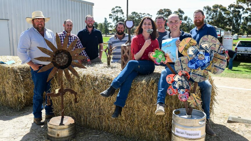 Woman with seven men sitting on and around hay bales with metal flowers. All smiling. 