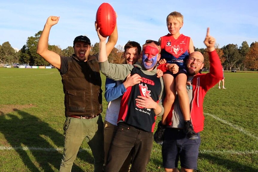 Four men and a boy wearing Demons club colours (blue and red) cheer and point on the football field.