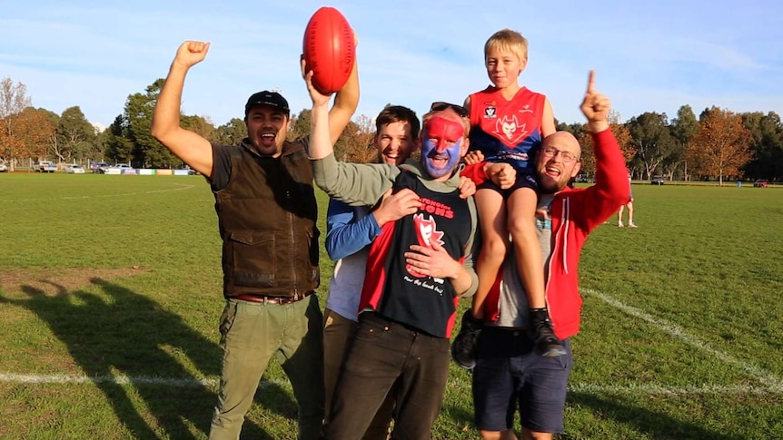 Four men and a boy wearing Demons club colours (blue and red) cheer and point on the football field.