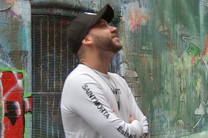 A man in a black cap and white long sleeve top stands in a laneway with graffiti covered walls looking up at a wall.