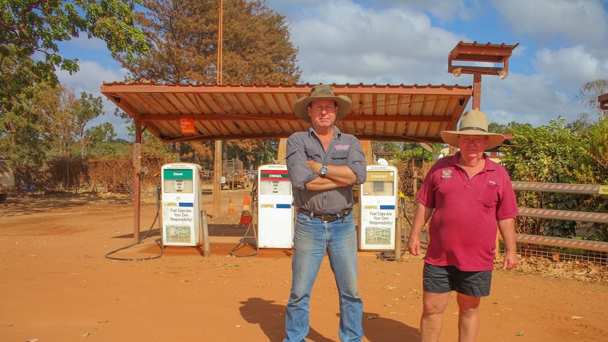 Two men in hats stand in the red dirt outside an ancient-looking petrol station.