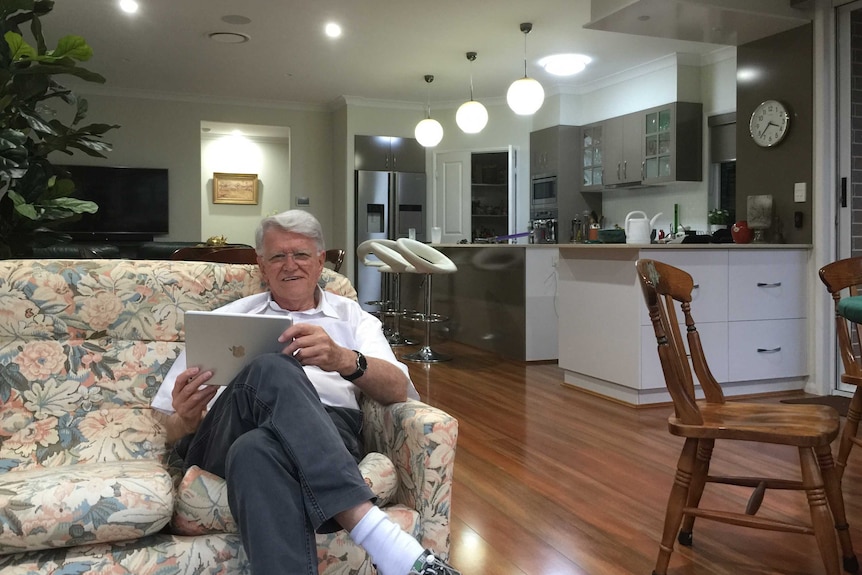 A man in his eighties sits on a couch in his living room