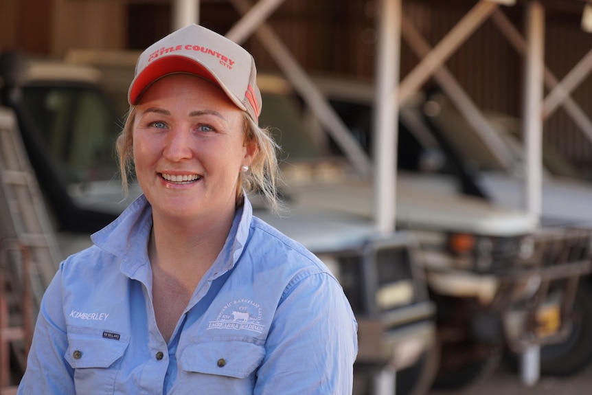 A woman in a blue shirt and cap with utes in the background.