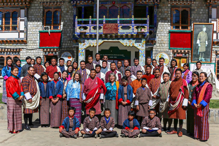 A group of teachers and students in Bhutan wearing brightly coloured national clothing stand in front of a school building.