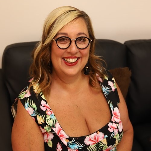 A woman with floral dress, black-rimmed glasses and fair hair sits on a couch and smiles widely.