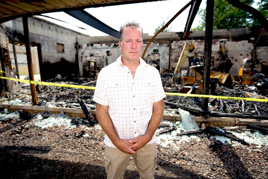 A man stands in front of a burned out building surrounded by police tape