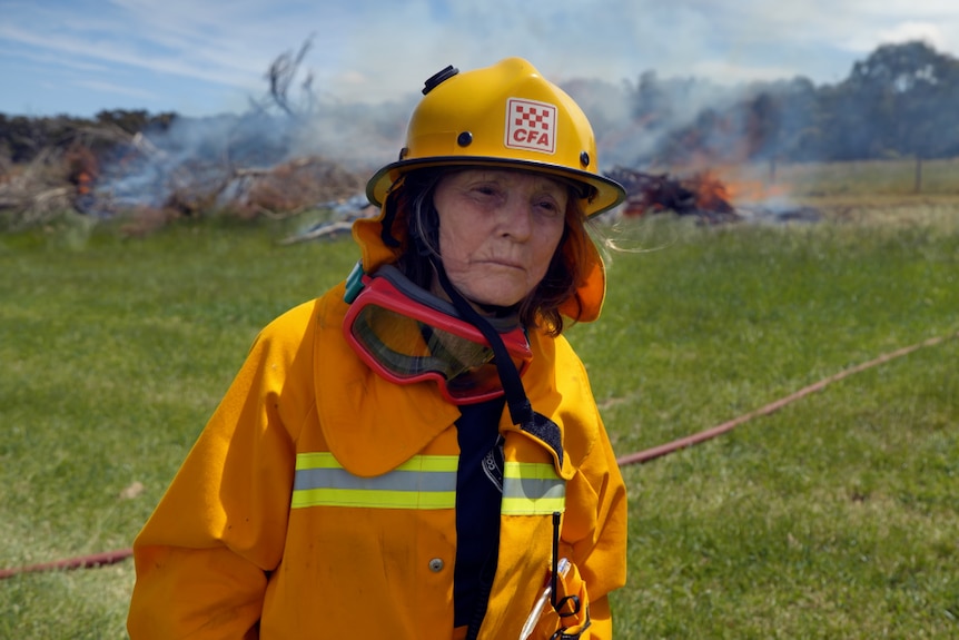 Woman wearing CFA uniform, goggles and hard hat standing in a field with a fire in the background. 