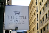 A white and blue sign reading The Little Hunter Steak & Ale Bar. In the background is a yellow building