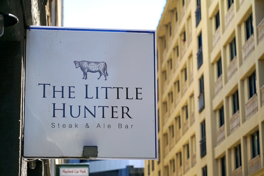 A white and blue sign reading The Little Hunter Steak & Ale Bar. In the background is a yellow building