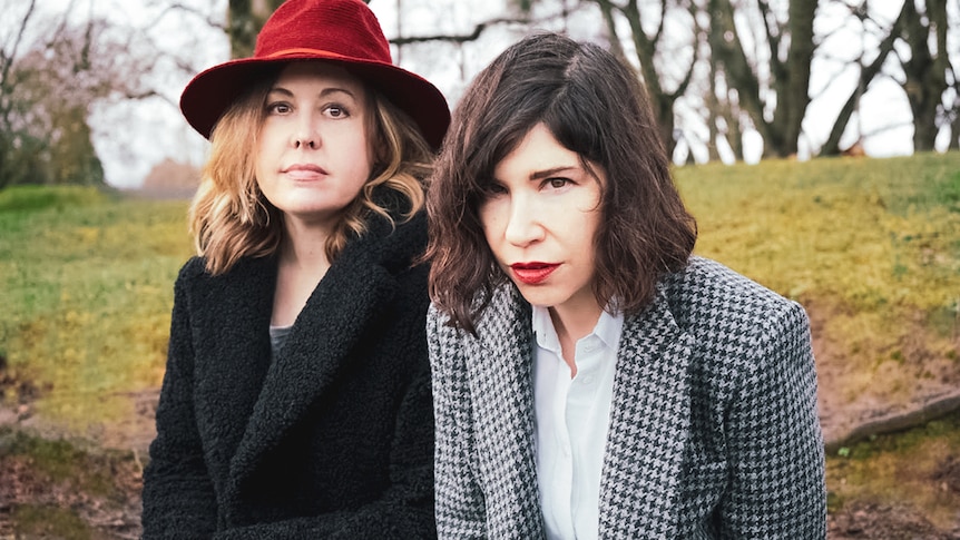 Corin Tucker and Carrie Brownstein of Sleater-Kinney sit in an empty park wearing coats. Corin also wears a red hat.