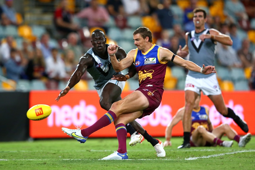 An AFL full-forward grimaces as he kicks the ball for a goal while a defender dives to stop him.