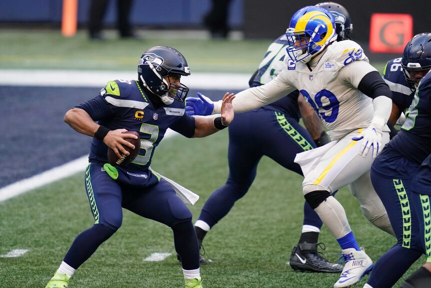 Russell Wilson holds an American football in one hand and fends at a player in white and blue kit who is running to him