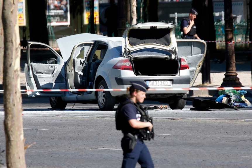 Armed police patrol an area where a white car is seen behind police tape.