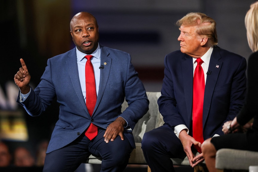 Tim Scott holds his hand up as Trump sits next to him looking at him