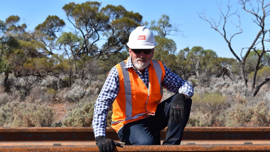 A man wearing sunglasses and a hard hat kneel down on a rail road.  His hand is on the rail