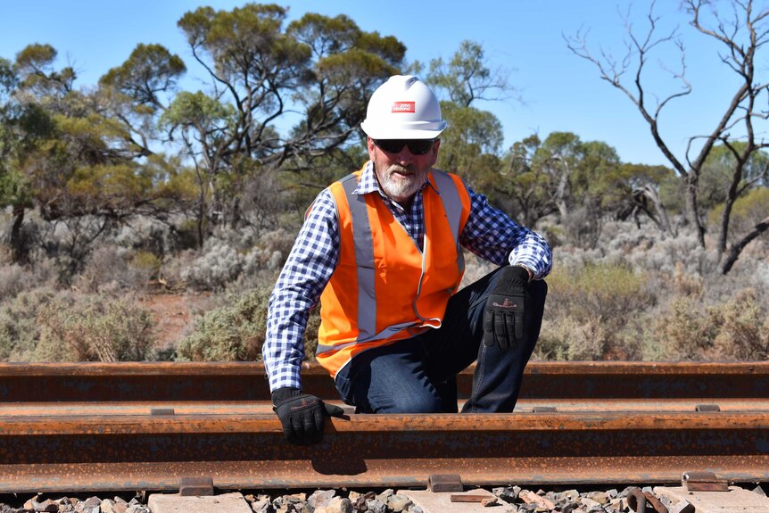 A man wearing sunglasses and a hard hat kneel down on a rail road.  His hand is on the rail