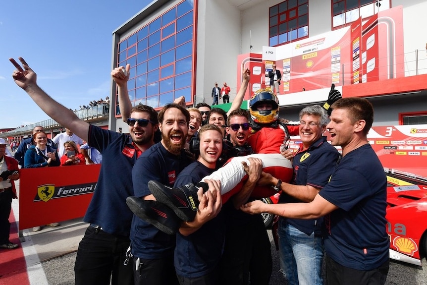 Woman race car driver in overalls and helmet being held up by team of men who are all cheering at a race track.