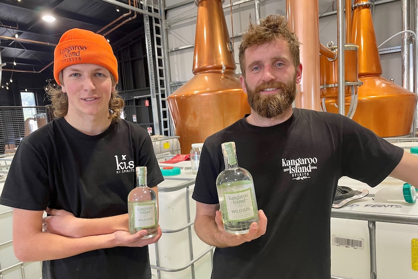 Two men smile as they hold bottles of gin in a distillery room.