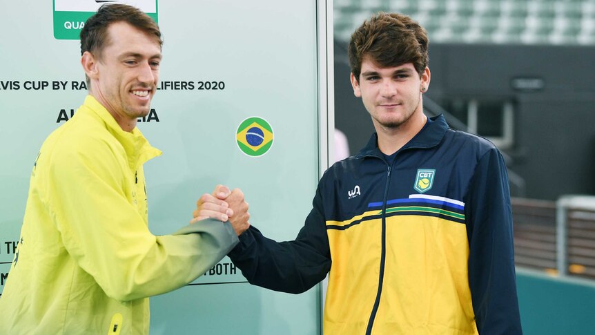 Australian and Brazilian tennis players shake hands in front of board showing a Davis Cup tie draw.