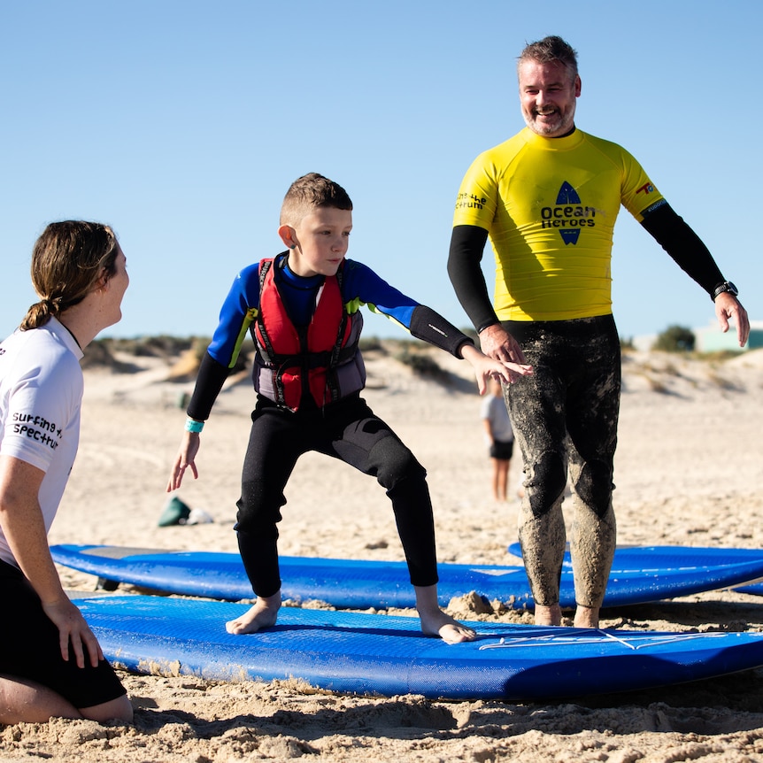 A child in a red life jacket and wetsuit stands on a surfboard on sand. Two adults instruct him.