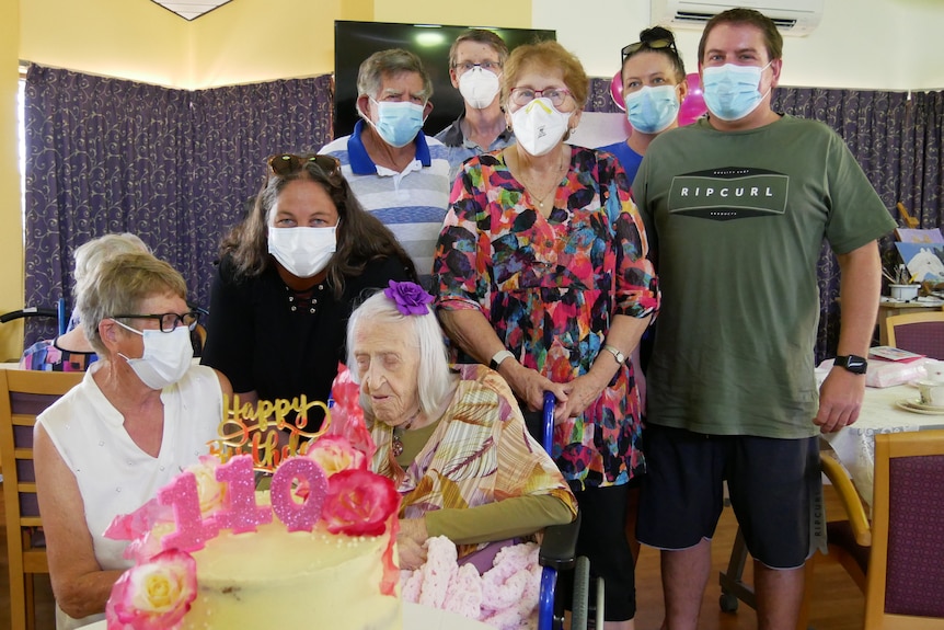 A photo of Dulcie Fawcett sitting in front of a birthday cake, surrounded by family and friends.