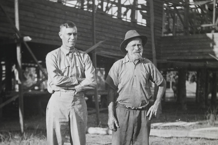 A black and white image of two men standing in front of wooden church under construction. 