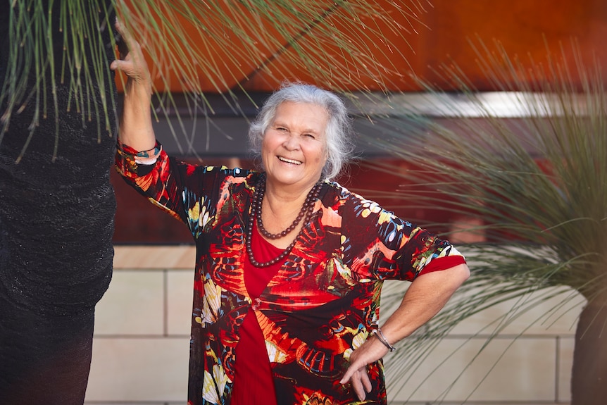 A 78-year-old First Nations woman smiles brightly. She has one arm on the trunk of a tree, the other on her hip.