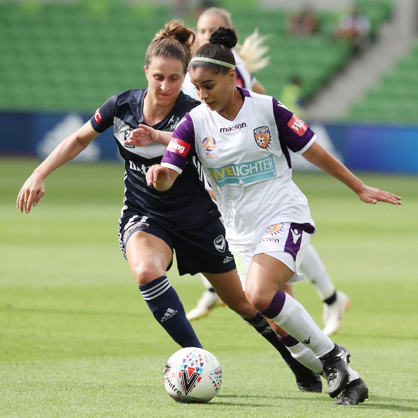 A women in a white kit dribbles the ball in front of a woman in a dark blue kit