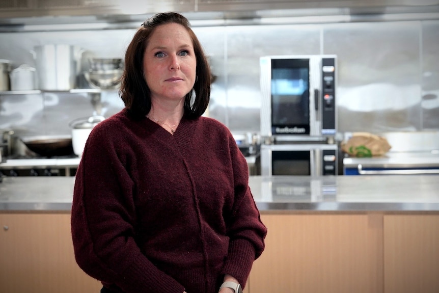 A middle-aged woman with a dark red top and dark hair stands in a kitchen posing for a photo with a serious expression.