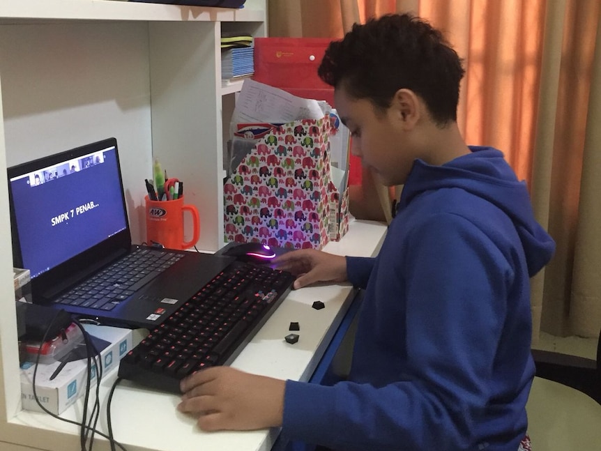 A young, dark-haired Indonesian student has logged on for his class on a laptop. He's wearing a royal blue hoodie.