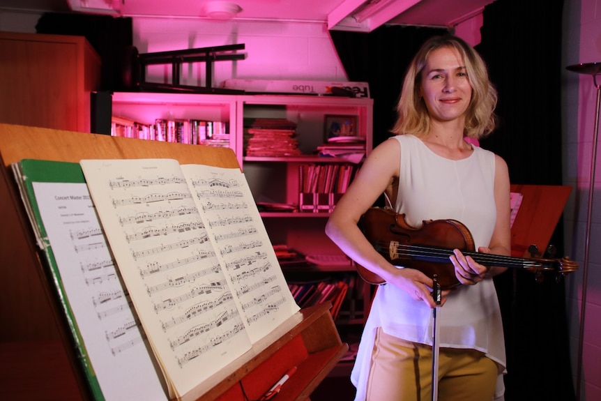 A woman poses for a photo holding a violin. There is a music stand in the foreground