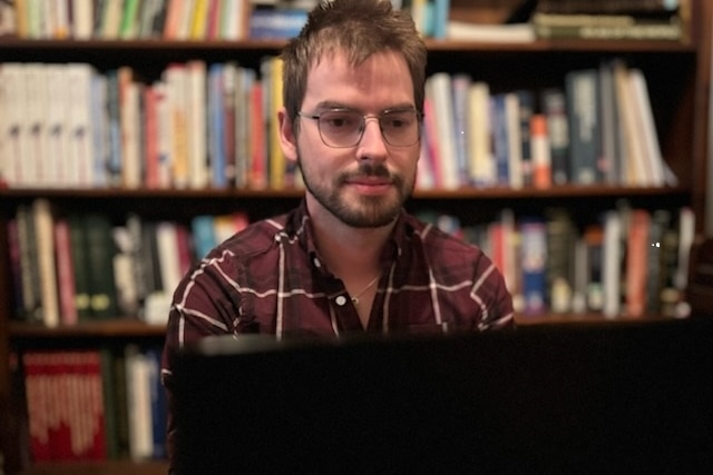 A young man facing the computer screen with a bookshelf behind him