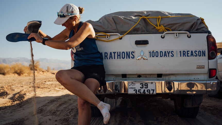 On a sandy road, a woman leans on utility vehicle trailer and pours sand from her running shoes.