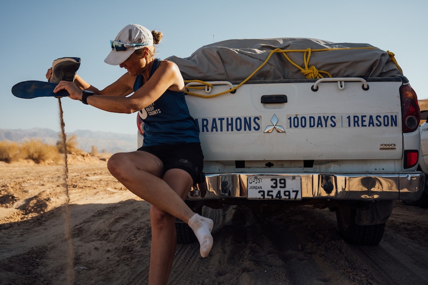 On a sandy road, a woman leans on utility vehicle trailer and pours sand from her running shoes.