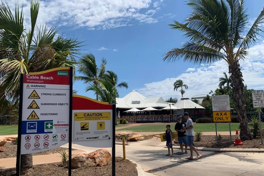 Three people stand near a sign warning of hazards at Cable Beach in Broome.