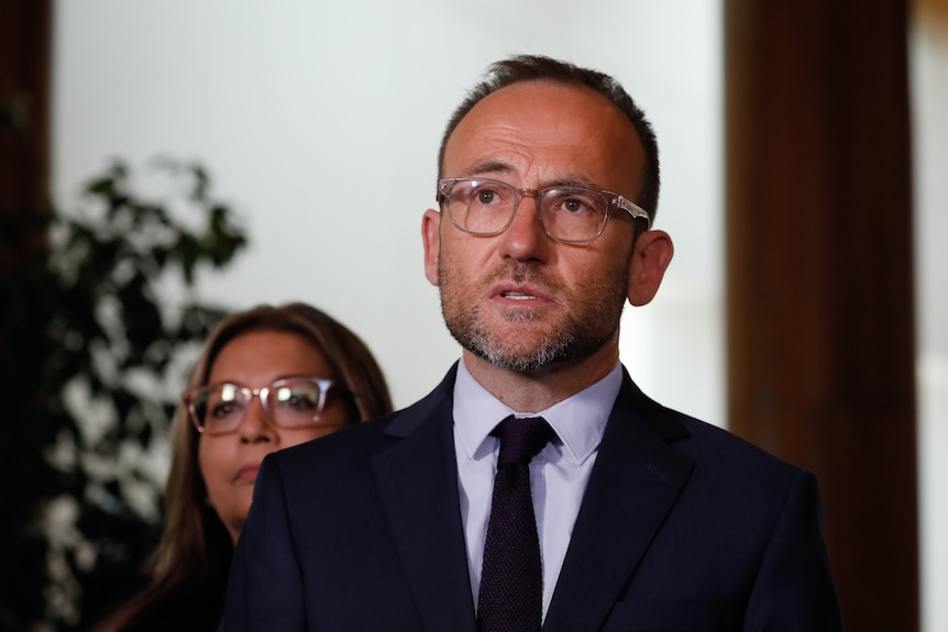 Adam Bandt wearing a suit and tie with the background out of focus, he's speaking and looking up to the top left of the photo