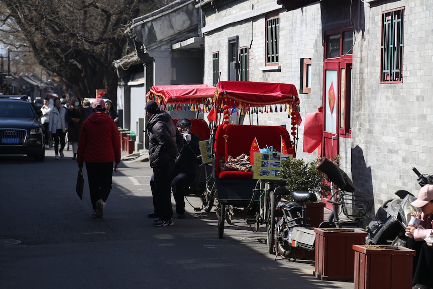 A man walk past a rickshaw driver set up near to rickshaws in a small alley.