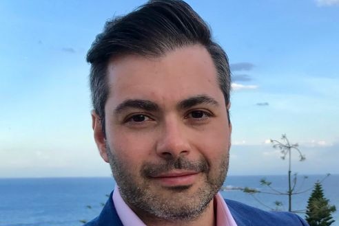 Close-up head shot of a man with a beard with the ocean behind him.