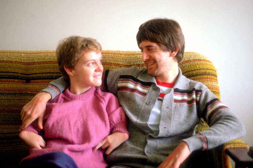 Lisa McManus and Andrew McManus, as a young couple, sit together on a couch looking at eachother fondly.