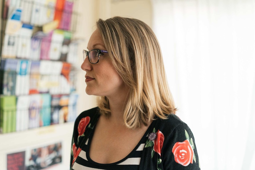 A woman with shoulder-length blonde hair, rose-print shirt and neutral expression stands in front of a wall of brochures.