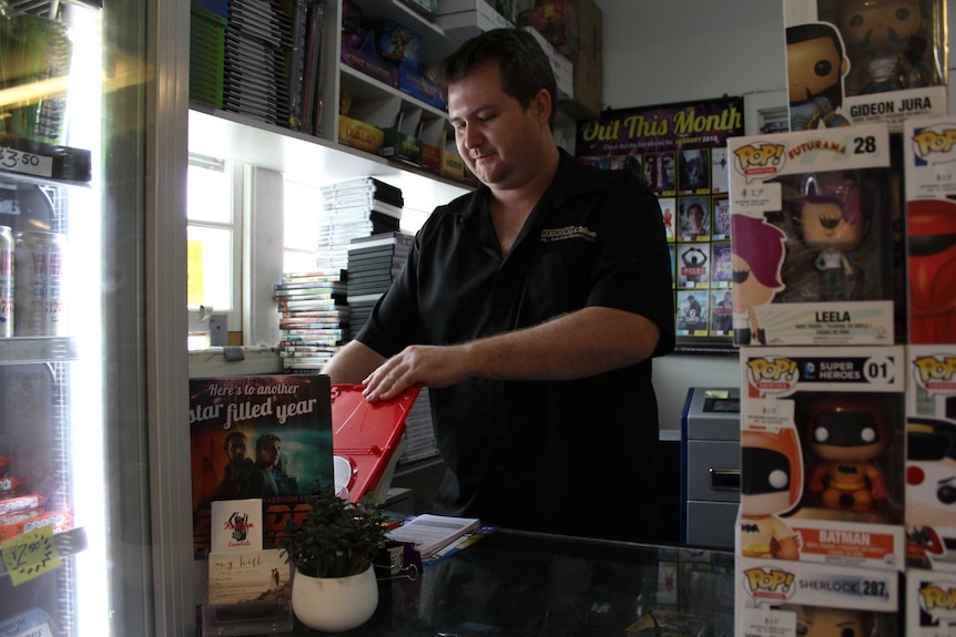Brenton Snell standing at the counter of his video store, scanning a DVD in a red plastic case.