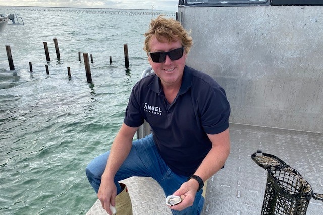 A smiling man in a blue tshirt holds an oyster while kneeling on a platform