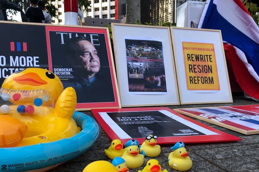 Rubber ducks on the ground in front of pictures, protest signs and the Thai flag.
