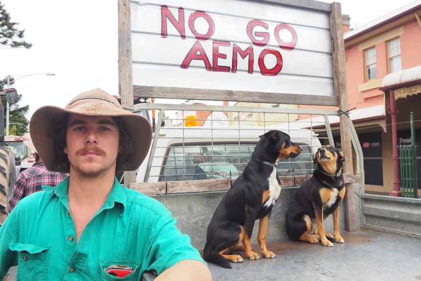 Farmer in a green shirt and brown had leans against a ute displaying a sign that reads: No go AEMO.